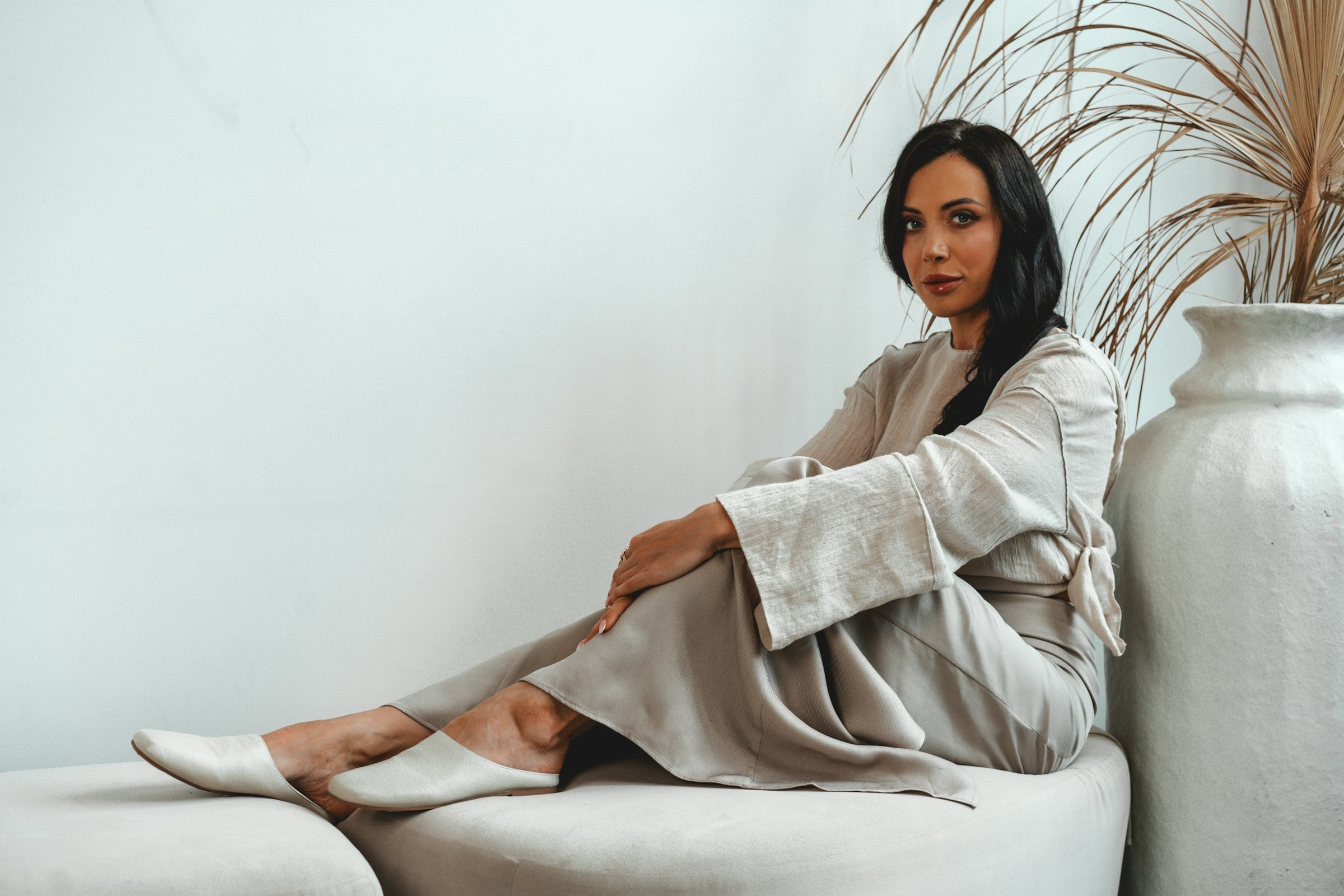 Woman in an ivory linen suit seated by a dried palm and a pale ceramic vase